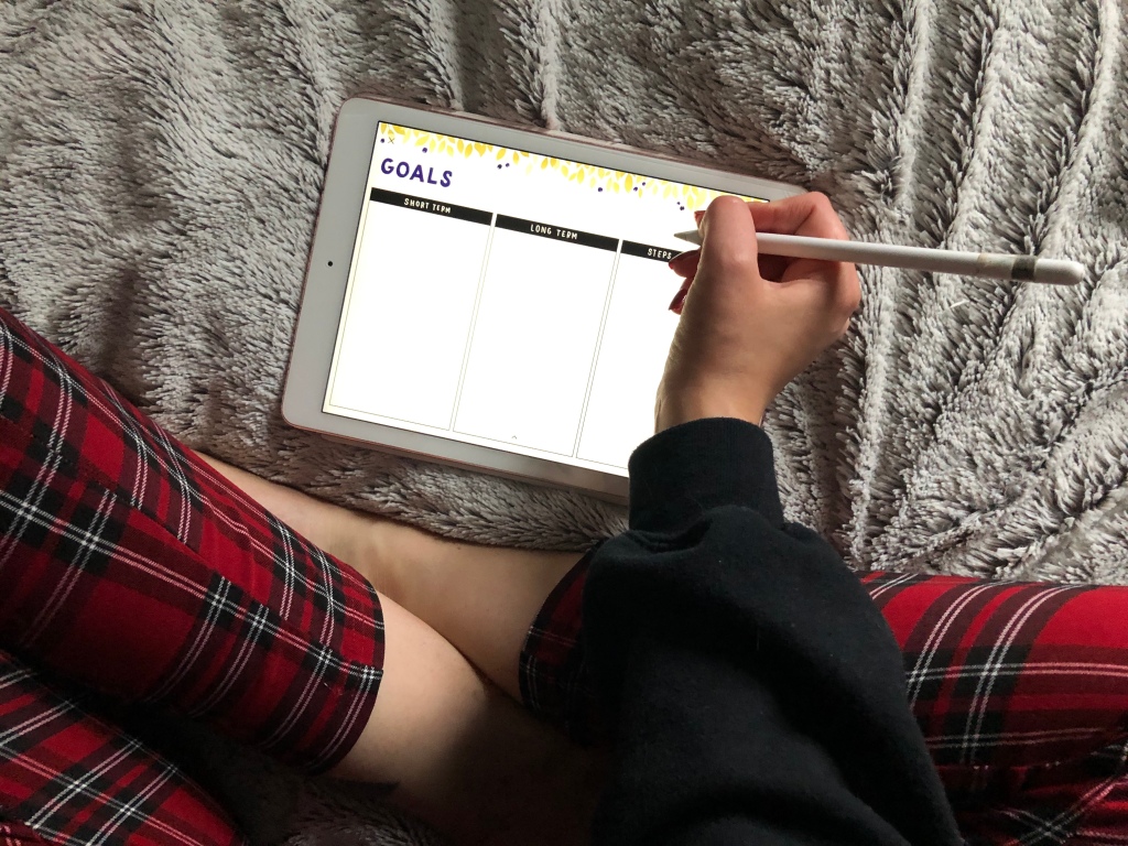A girl is wearing tartan trousers and writing on her iPad on a note book which reads 'Goals'